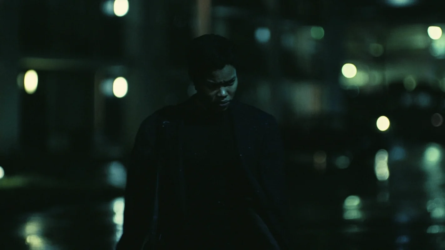 A man walks through a rain-darkened street beneath green city lights.