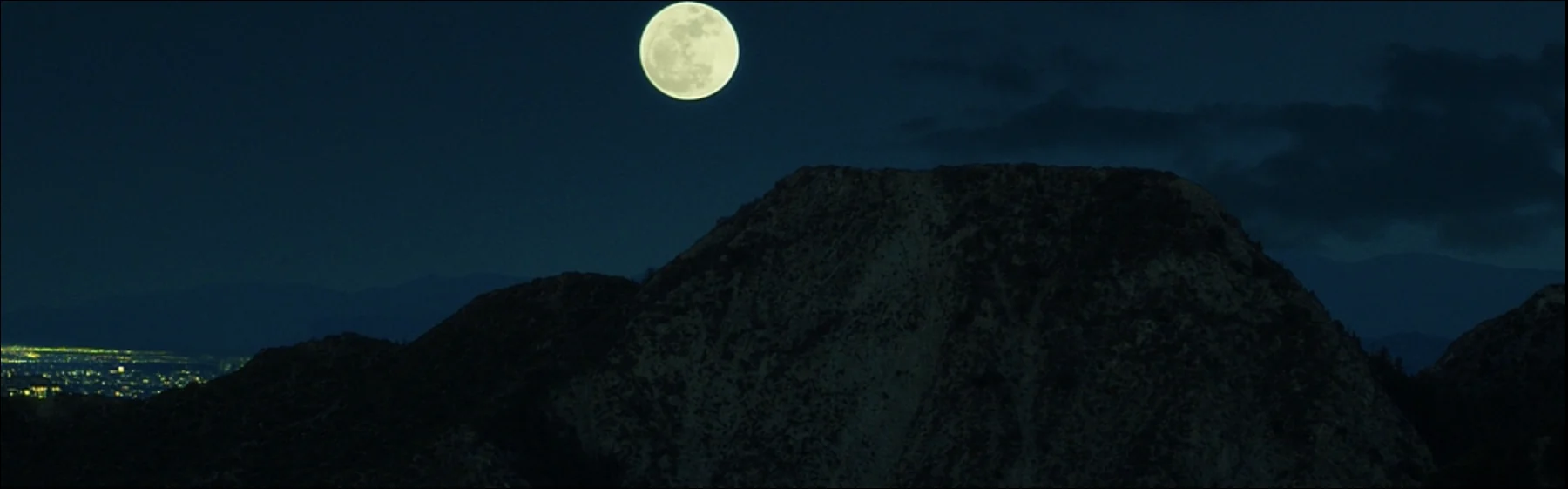 A full moon rises above a dark mountain ridge, with distant city lights.