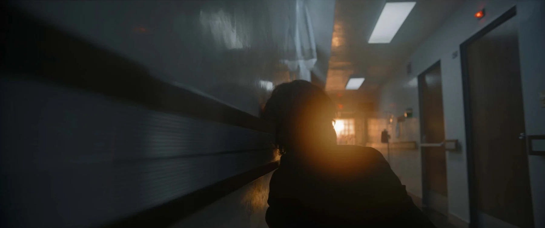 A slumped figure in a hospital corridor is silhouetted against low sunlight.