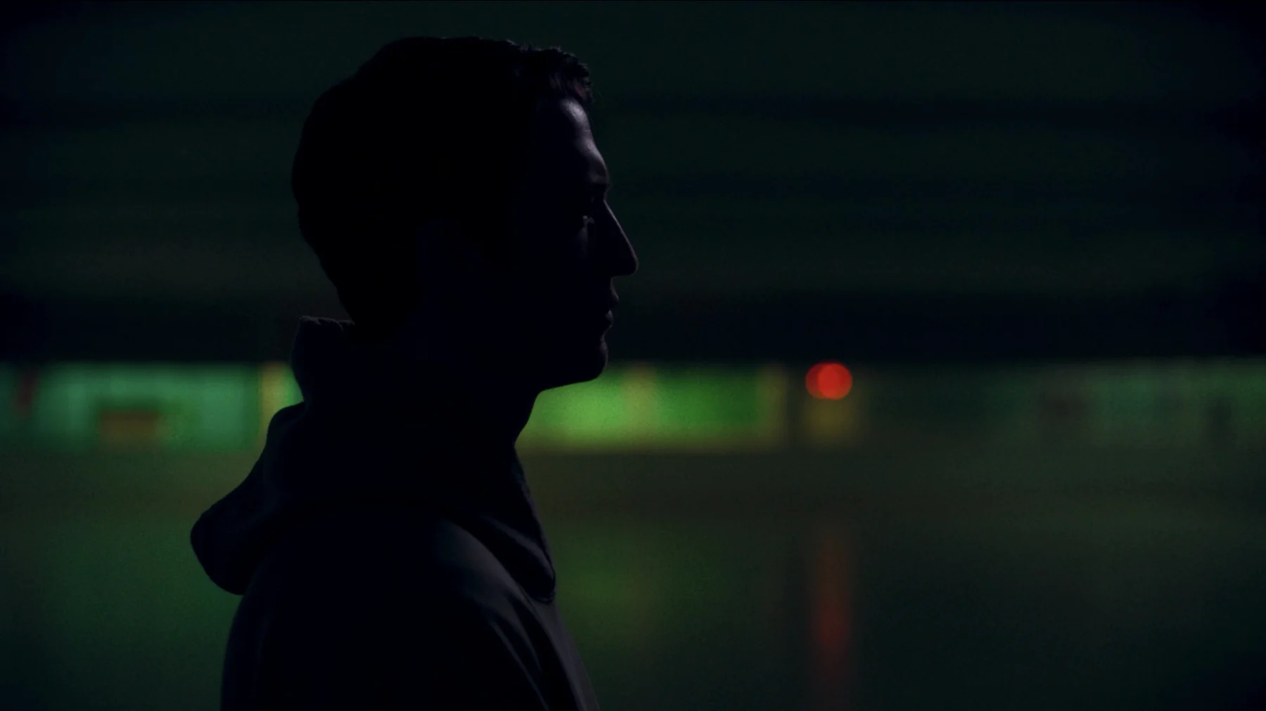 A man’s profile is silhouetted against green light and a distant red signal.