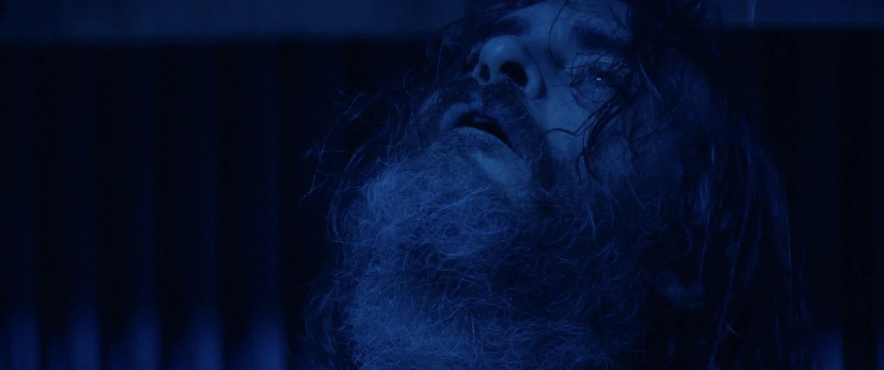A bearded man looks upward in deep blue light.