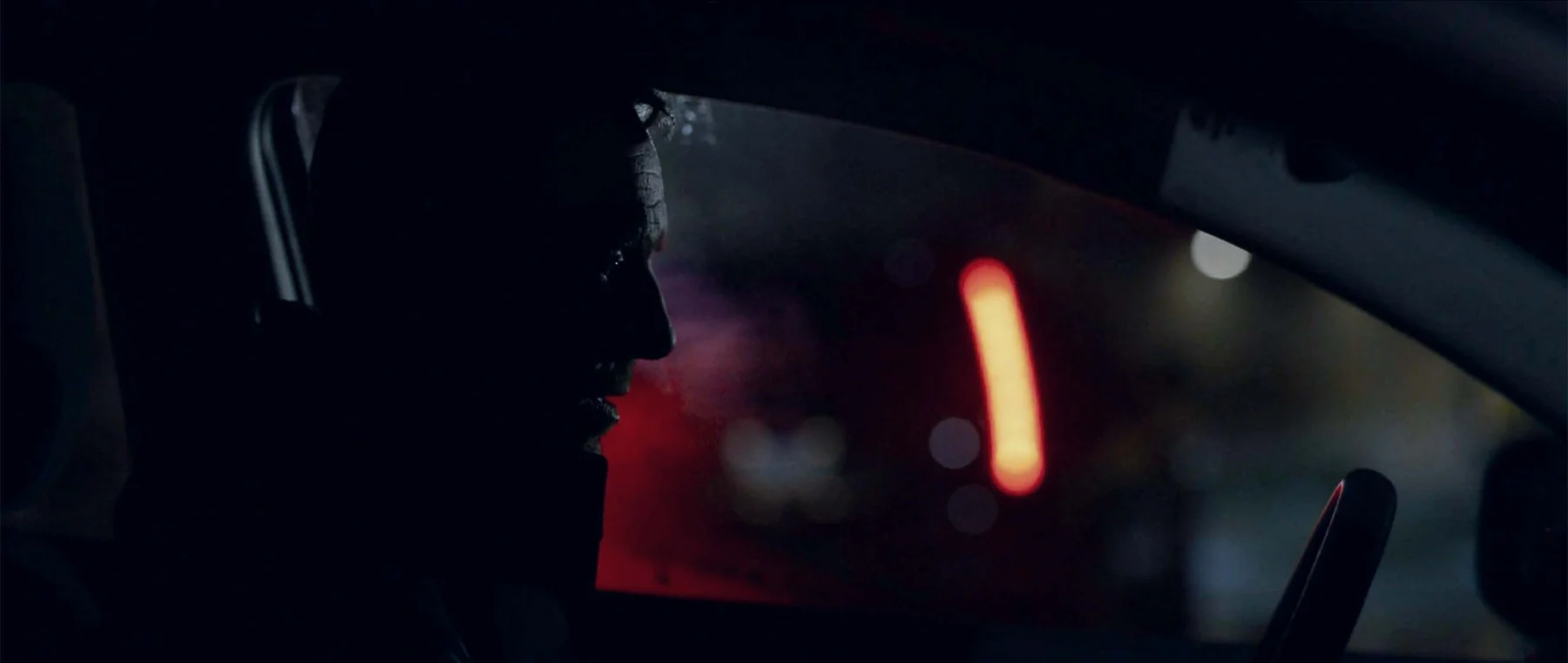 A driver’s profile is silhouetted against a red light outside the car.