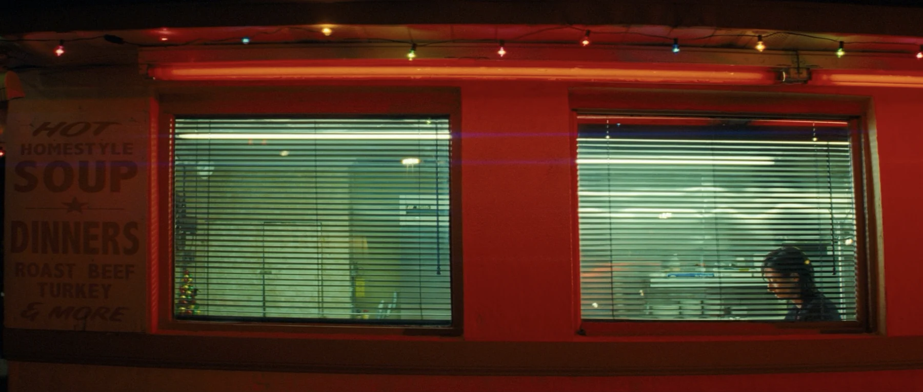 A woman sits behind the blinds of a red-lit diner at night.