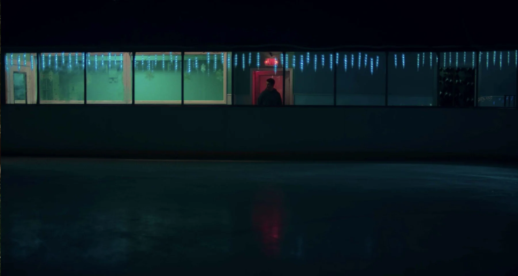A lone man sits within the wide diner windows and red doorway at night.
