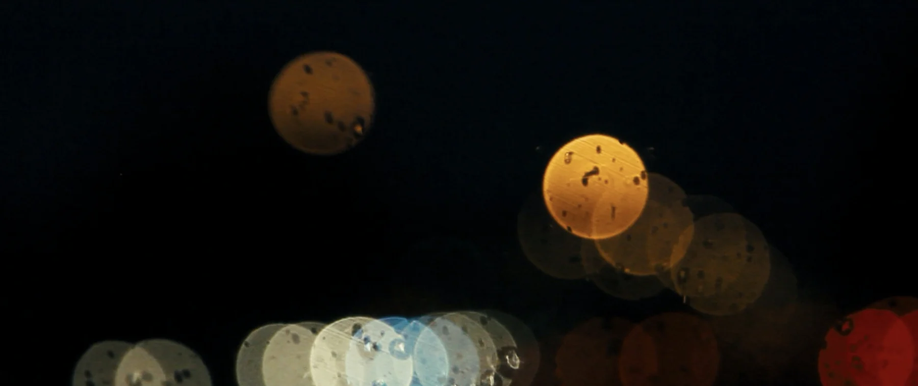 Rain-marked circles of amber, white, and red light overlap in darkness.