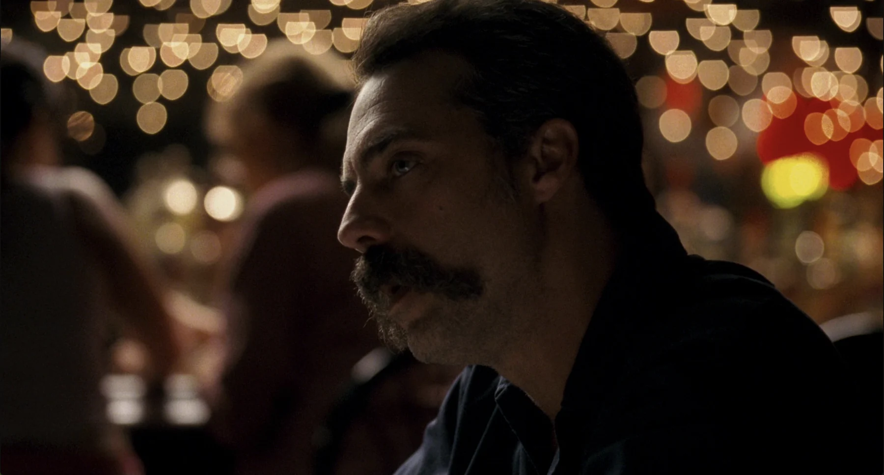 A bearded man sits in a crowded restaurant beneath warm lights.