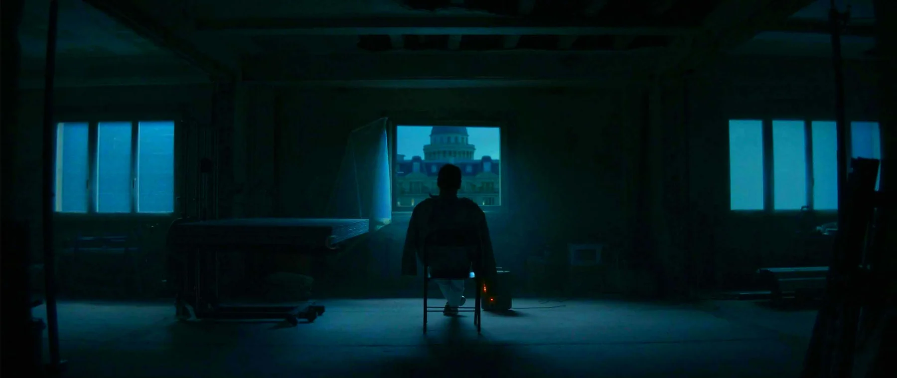 A solitary figure sits within the precise geometry of a blue-lit room.