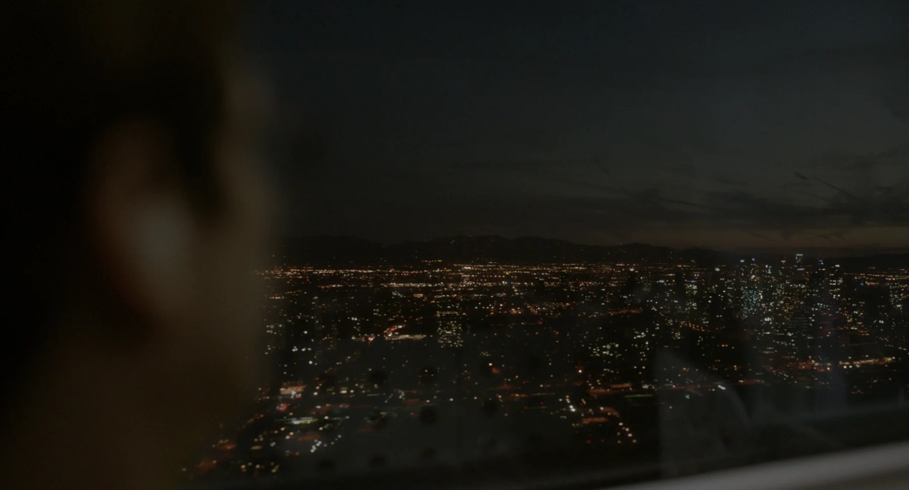 A blurred figure overlooks city lights through a window at dusk.