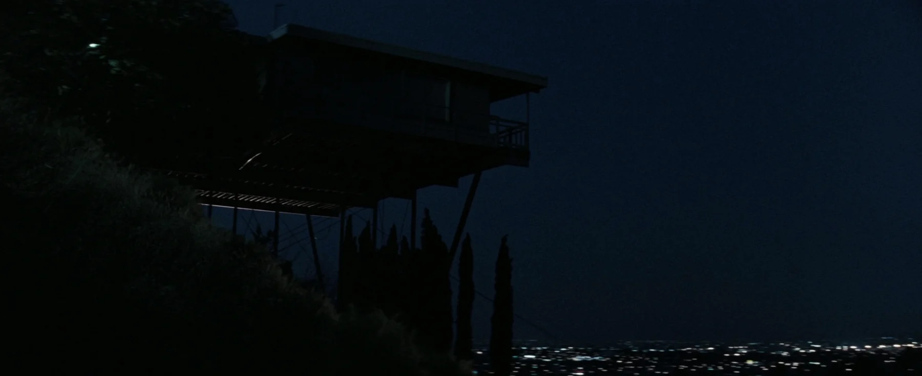A dark hillside house projects over a distant city lit beneath a blue night sky.